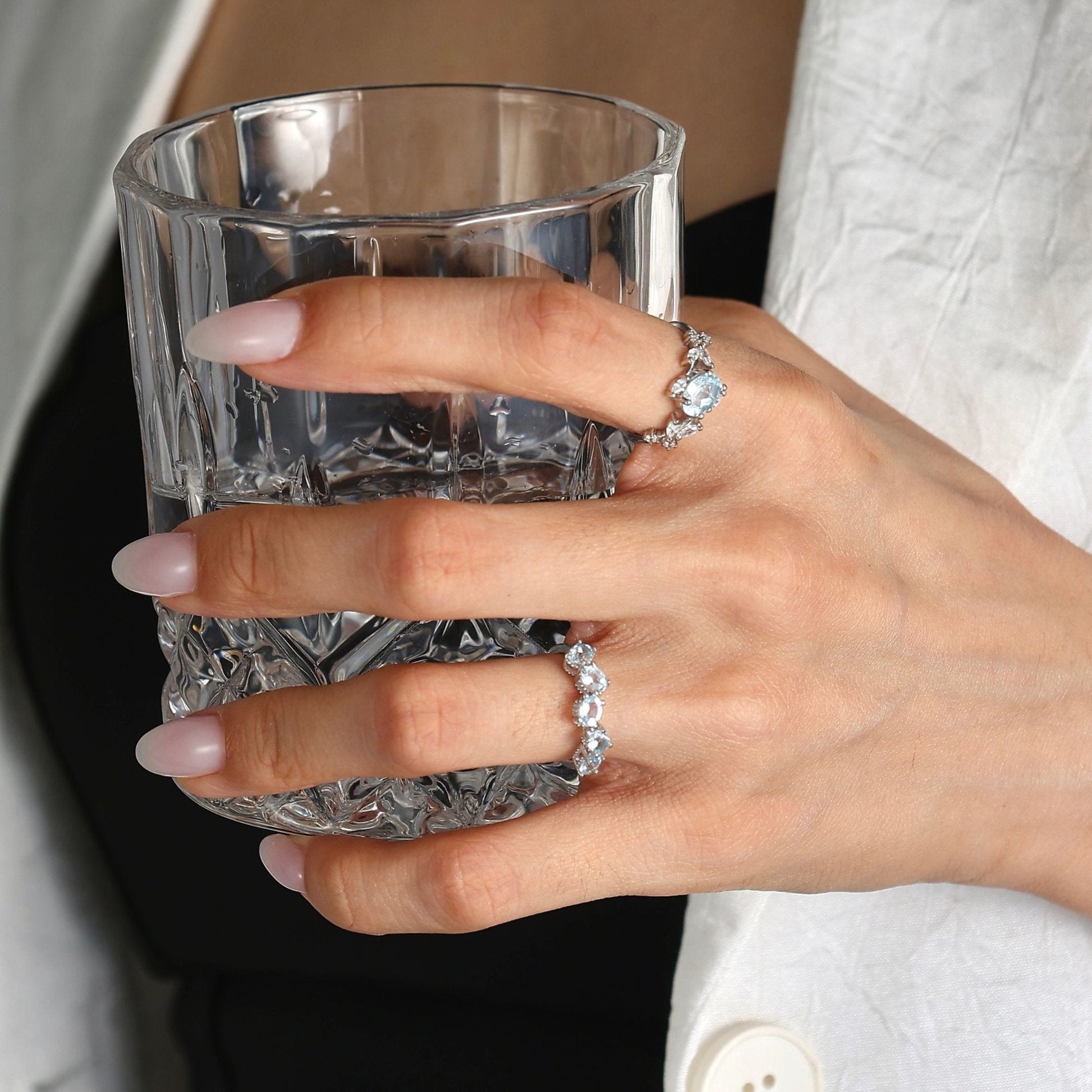 Lifestyle photograph of a woman's hand, wearing a white blazer and black top, holding a cut crystal glass. The focus is on her ring finger and middle finger, showcasing the Awakened Time sterling silver jewelry set: a central floral-inspired aquamarine ring paired with a matching clustered aquamarine eternity band, set against a soft, blurred background.