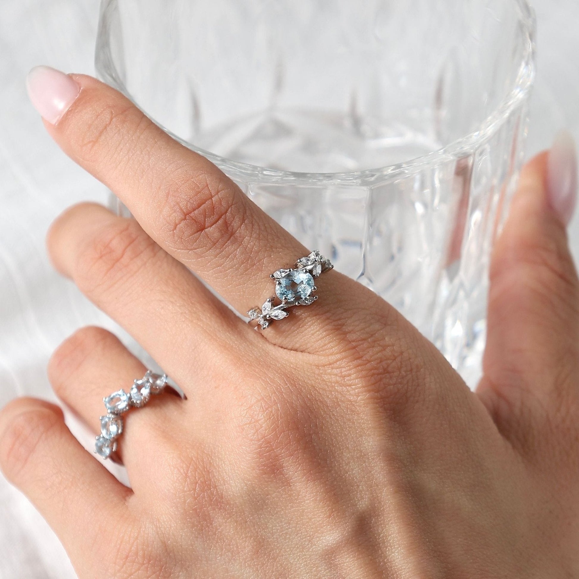 Lifestyle photograph of a woman wearing an Awakened Time sterling silver light blue aquamarine jewelry set while holding a clear glass, featuring a floral-inspired aquamarine ring on her finger and a matching stone-accented band on an adjacent finger against a light background.