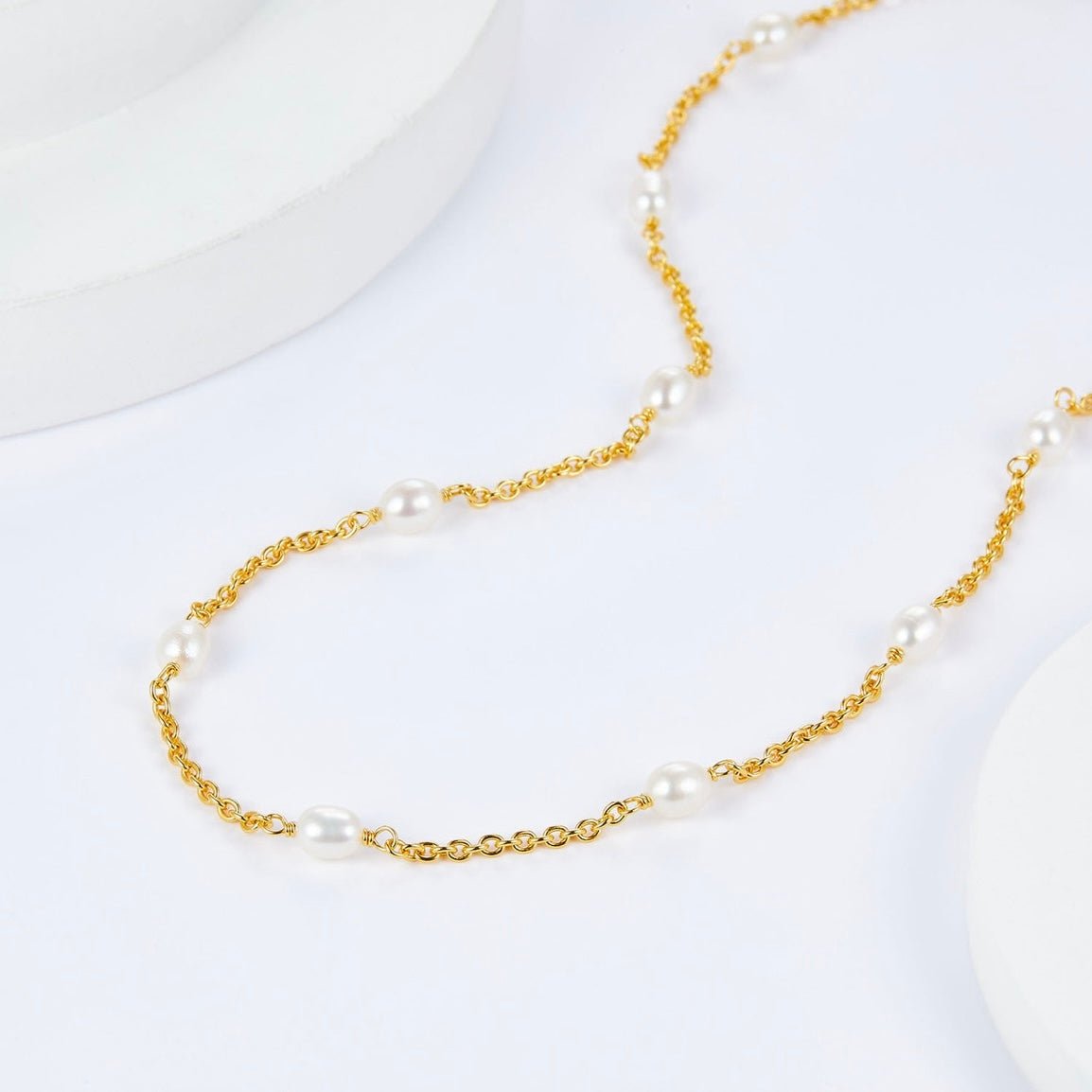 Studio view of the Awakened Time Bare Luxe Orbit gold pearl chain resting gracefully on a textured white surface.