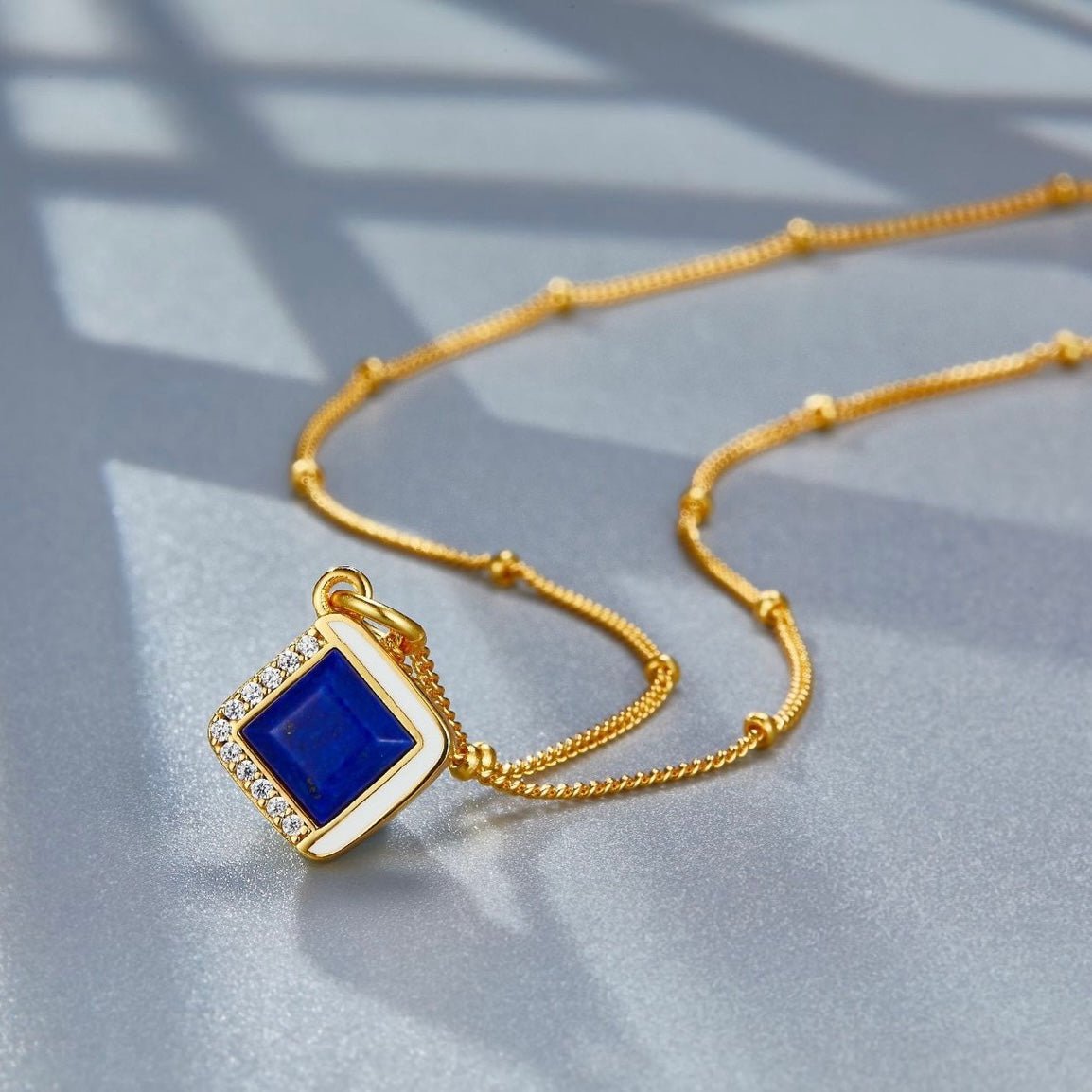 Studio view of the Awakened Time Bleu Intuition lapis lazuli pendant on a beaded gold chain resting on a shaded surface.