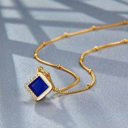 Studio view of the Awakened Time Bleu Intuition lapis lazuli pendant on a beaded gold chain resting on a shaded surface.