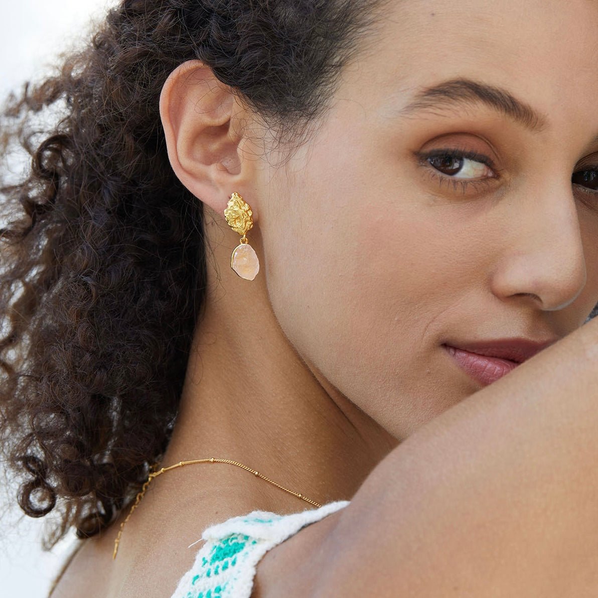 Model looking downward while wearing a green crochet top and Awakened Time raw rose quartz drop earrings.