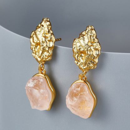 Both Awakened Time gold textured studs with raw rose quartz drops standing upright against a curved grey backdrop.