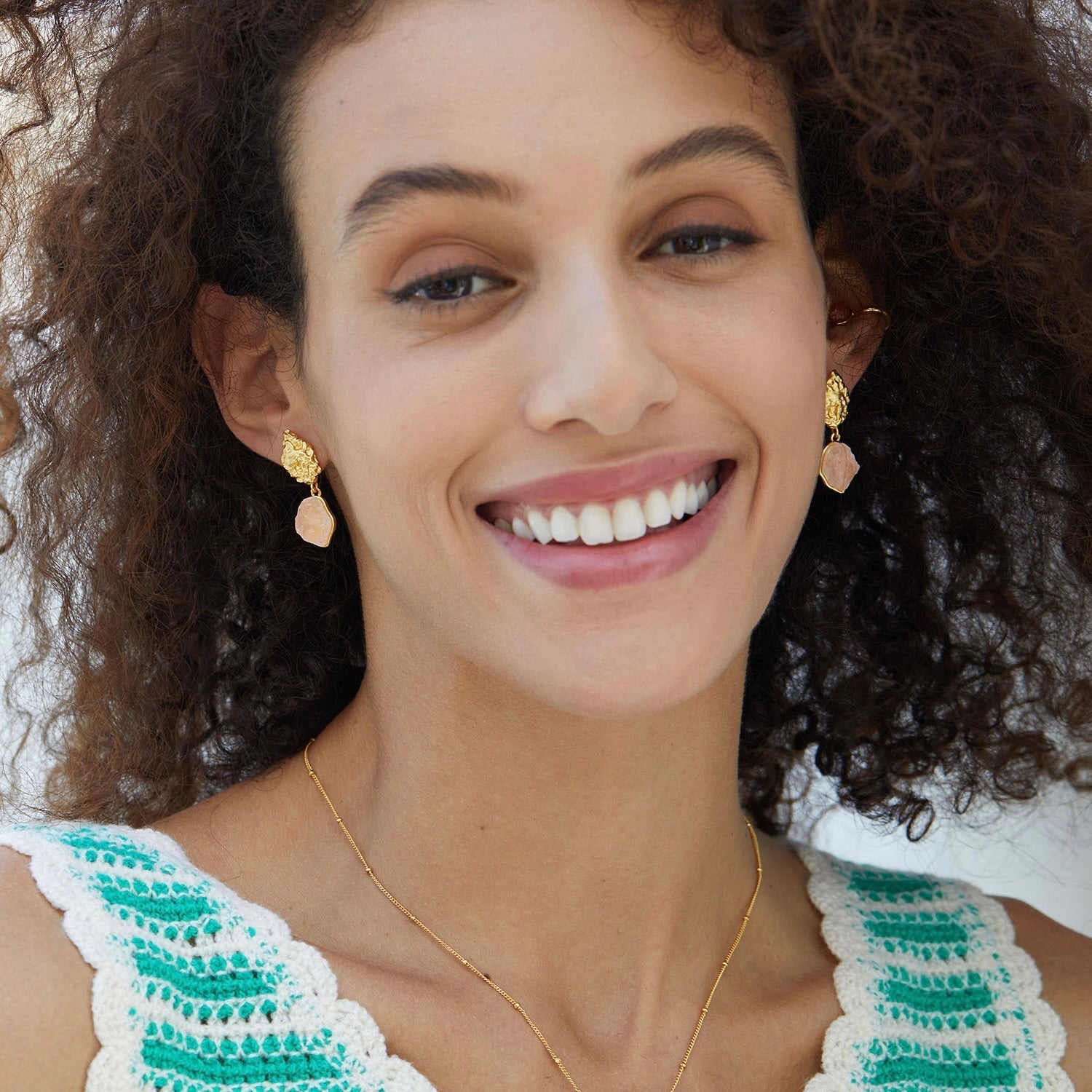 Smiling model showcasing Awakened Time gold textured stud earrings with raw pink rose quartz gemstone drops.