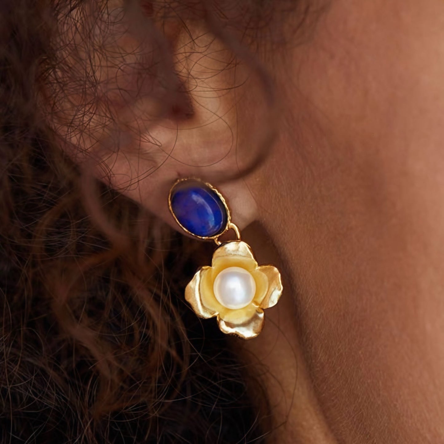 Macro close-up shot of a model wearing the Awakened Time Fleur de Lapis gold flower drop earring with deep blue lapis lazuli and freshwater pearl center.