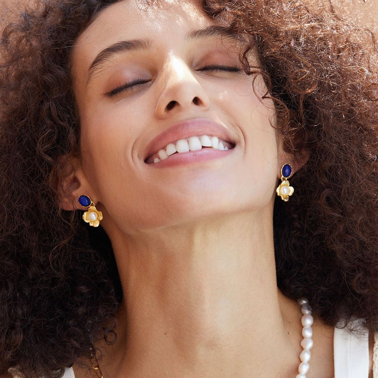 Model looking up joyfully wearing a pair of Awakened Time Fleur de Lapis gold floral drop earrings styled with a pearl necklace.