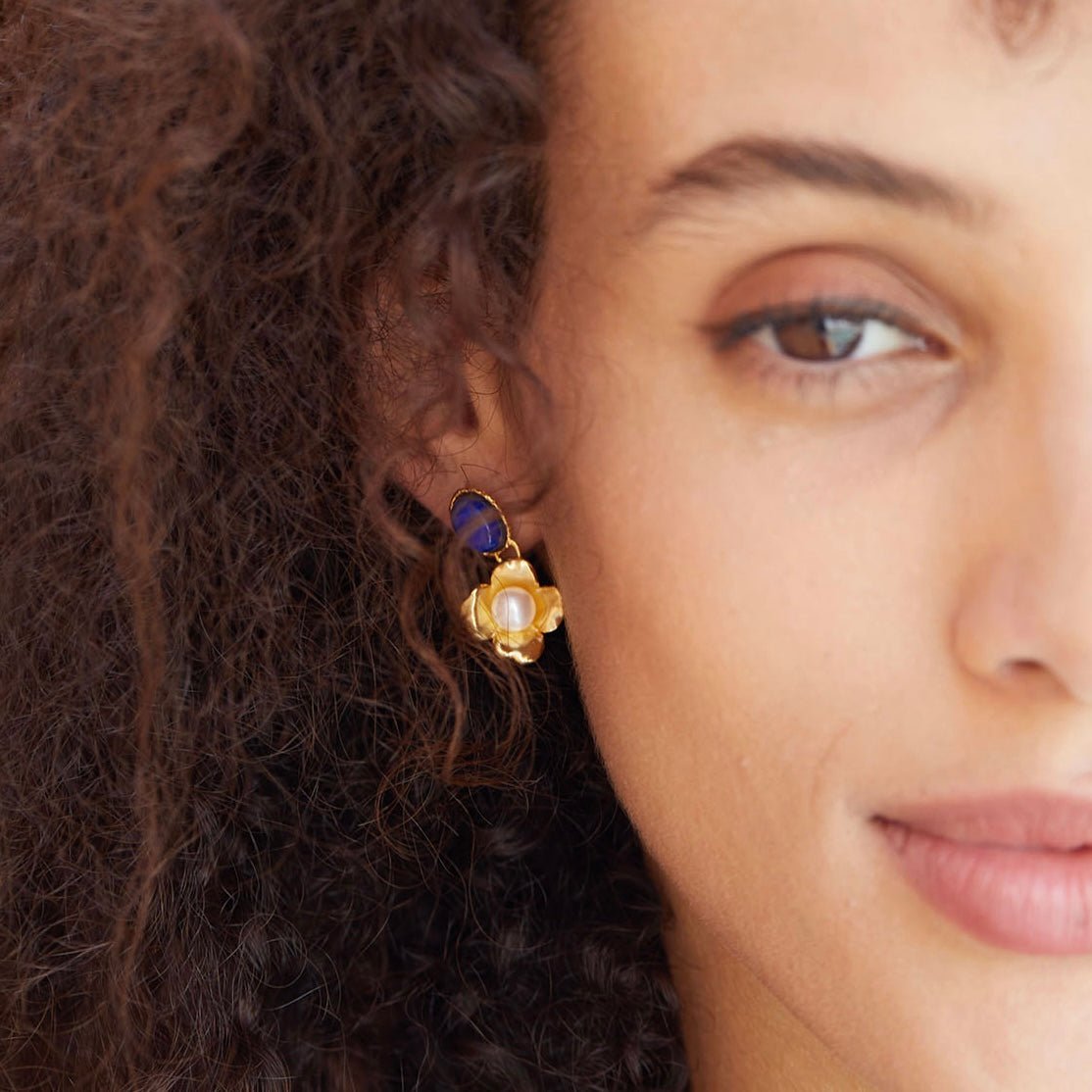 Model with curly brown hair wearing the Fleur de Lapis gold and blue lapis lazuli pearl earring shown from a front profile view.