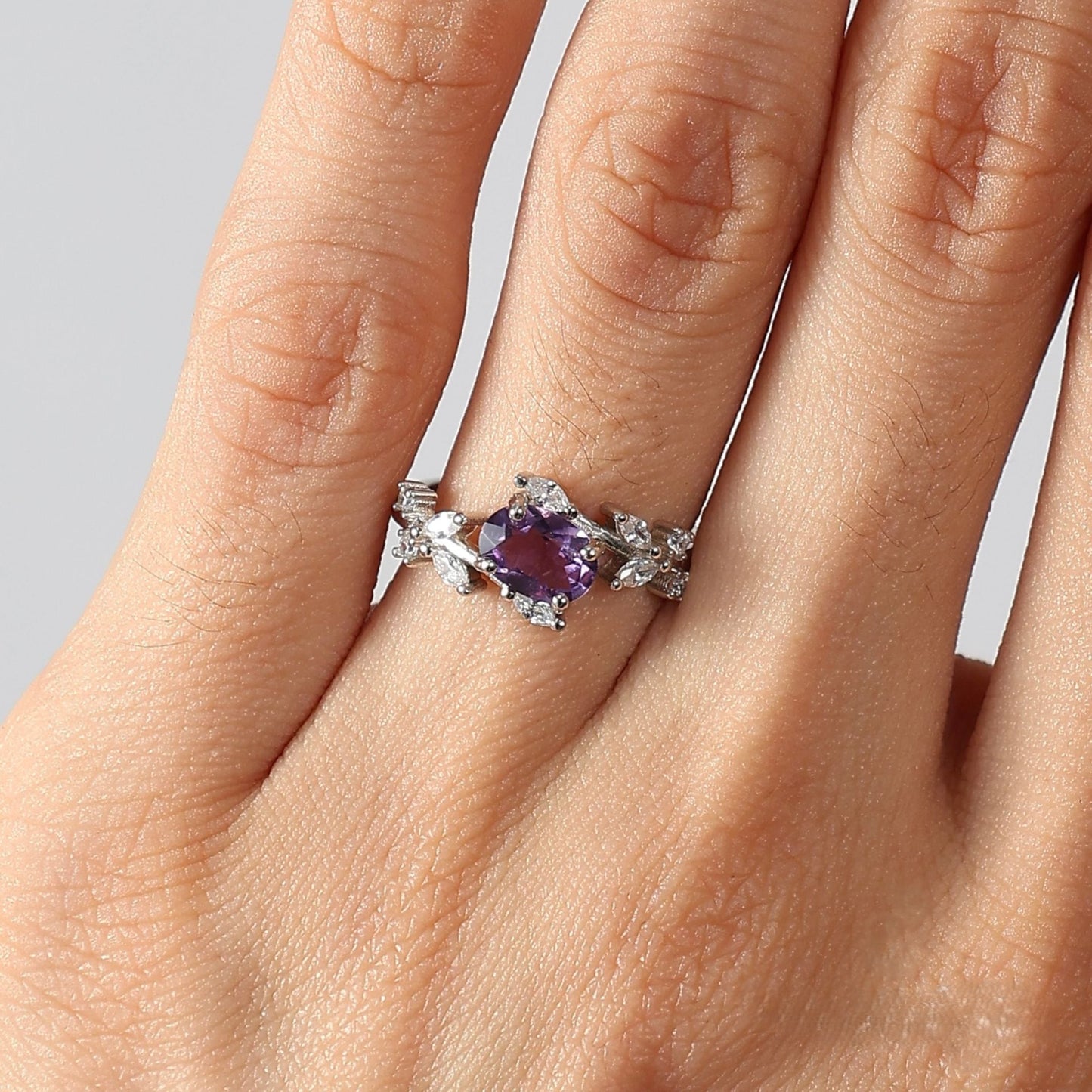 Close-up photograph of a woman's left hand, focusing on the ring finger wearing the Awakened Time Floral Ring. The ring features a central oval-cut natural amethyst set in 925 sterling silver, framed by a detailed vine-and-leaf motif adorned with white cubic zirconia, against a neutral studio background.