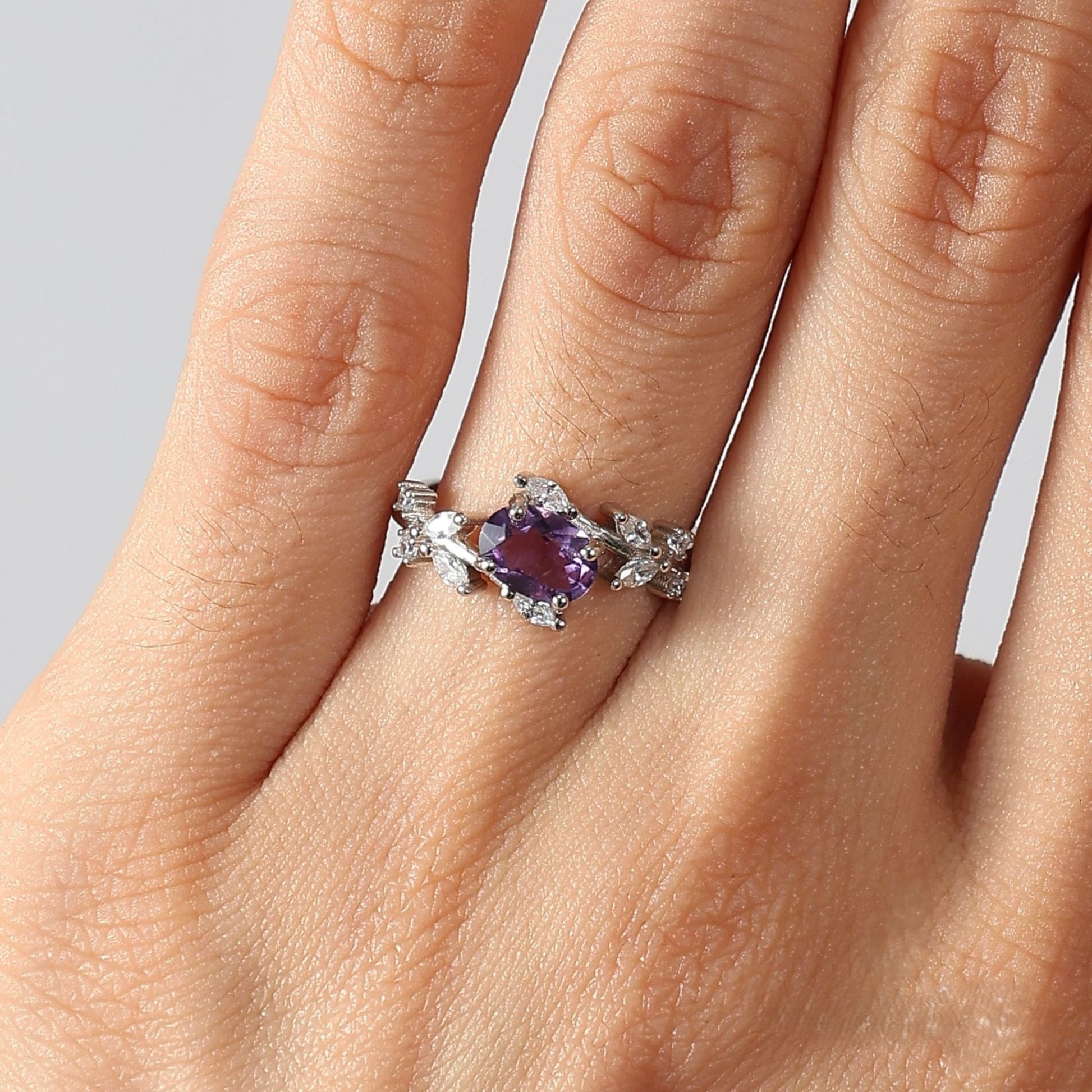 Close-up photograph of a woman's left hand, focusing on the ring finger wearing the Awakened Time Floral Ring. The ring features a central oval-cut natural amethyst set in 925 sterling silver, framed by a detailed vine-and-leaf motif adorned with white cubic zirconia, against a neutral studio background.