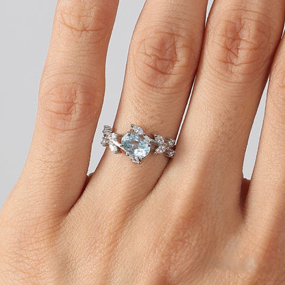 Close-up photograph of a woman's hand, focusing on the ring finger wearing the Awakened Time floral ring. The design features a central oval-cut light blue aquamarine set in 925 sterling silver, framed by a delicate vine-and-leaf motif adorned with white cubic zirconia against a neutral studio background.