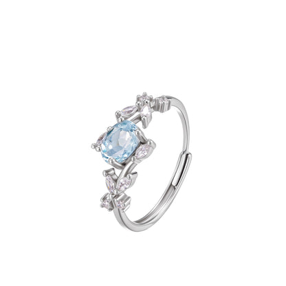 Close-up studio photograph of the AwakenedTime Floral Ring. The ring features an oval-cut natural light blue topaz gemstone set in 925 sterling silver, surrounded by a vine-and-leaf motif accented with sparkling cubic zirconia, shown on a white background.