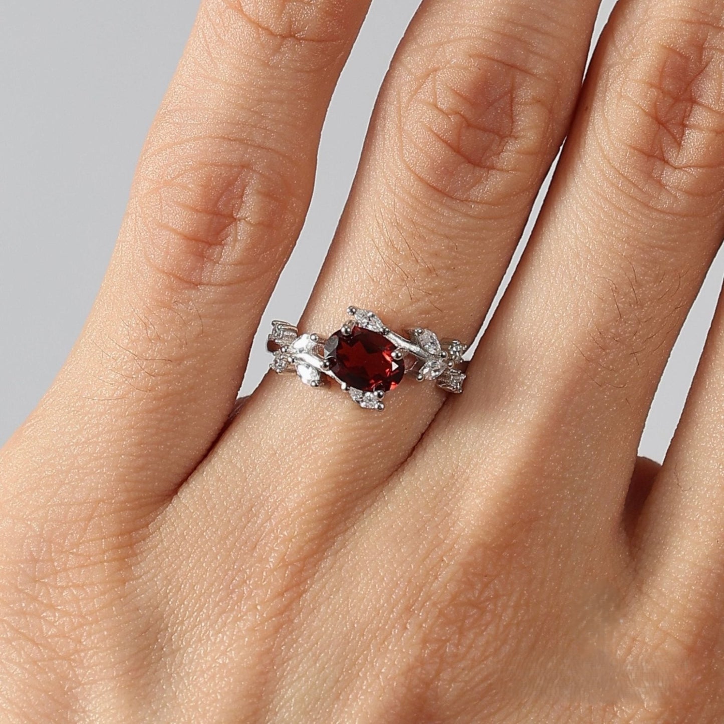 Close-up photograph of a woman's hand, focusing on the ring finger wearing the Awakened Time Floral Ring. The ring features a central oval-cut deep red garnet set in 925 sterling silver, framed by a detailed vine-and-leaf motif adorned with white cubic zirconia, against a neutral light grey studio background.