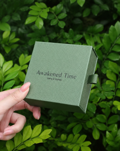Hand holding a sage green Awakened Time signature jewelry gift box surrounded by natural green foliage.