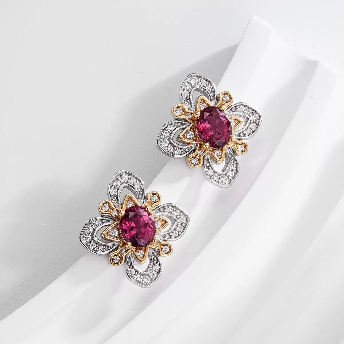 Pair of Awakened Time La Rosa di Firenze two-tone stud earrings displayed on a white textured pedestal, highlighting oval red garnet gemstones framed in gold petals and silver pavé zirconia.