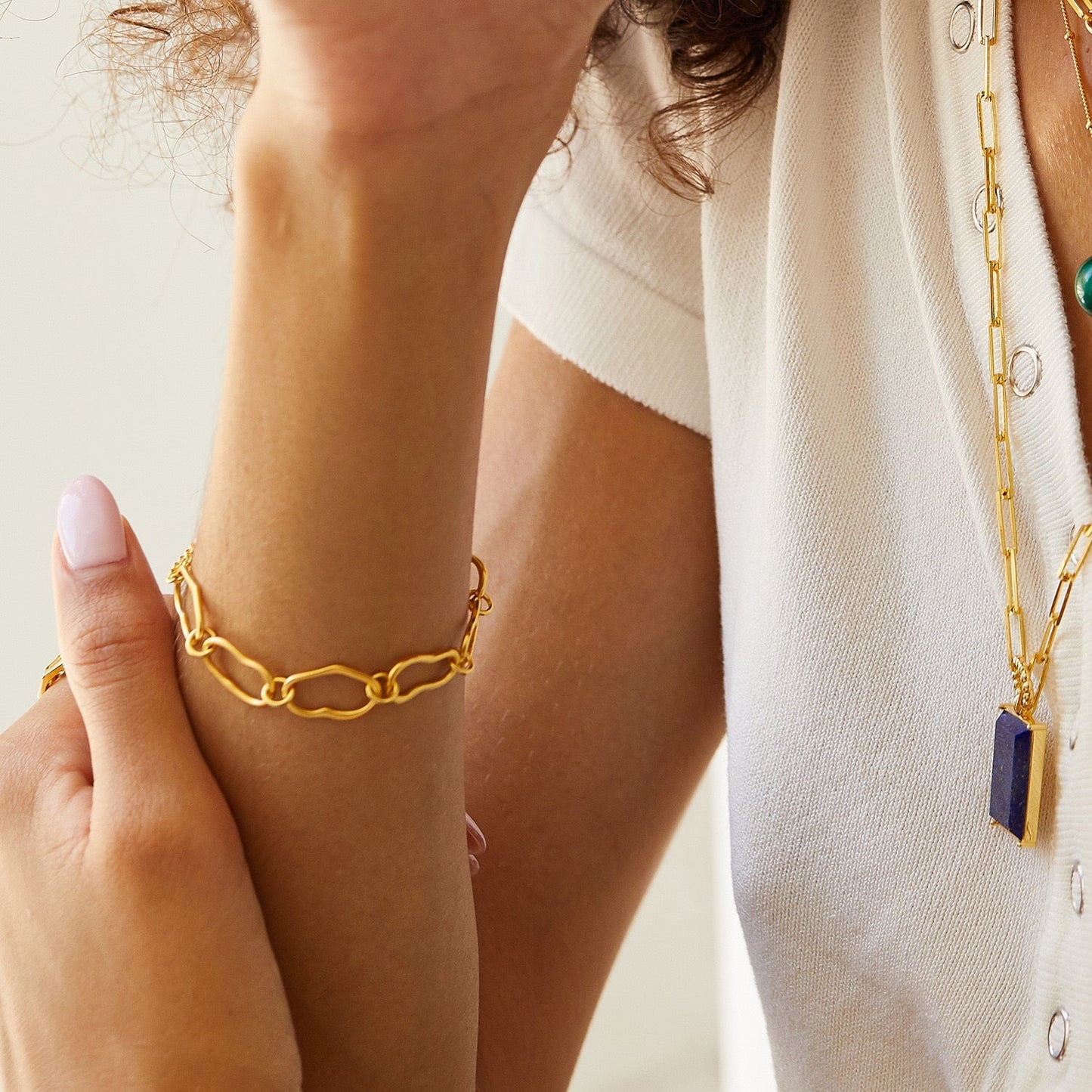 A close-up view of a woman wearing the Awakened Time Liaison gold chain bracelet, featuring interlocking irregular links. She also wears a matching gold long necklace with a blue lapis pendant and has a light pink manicure
