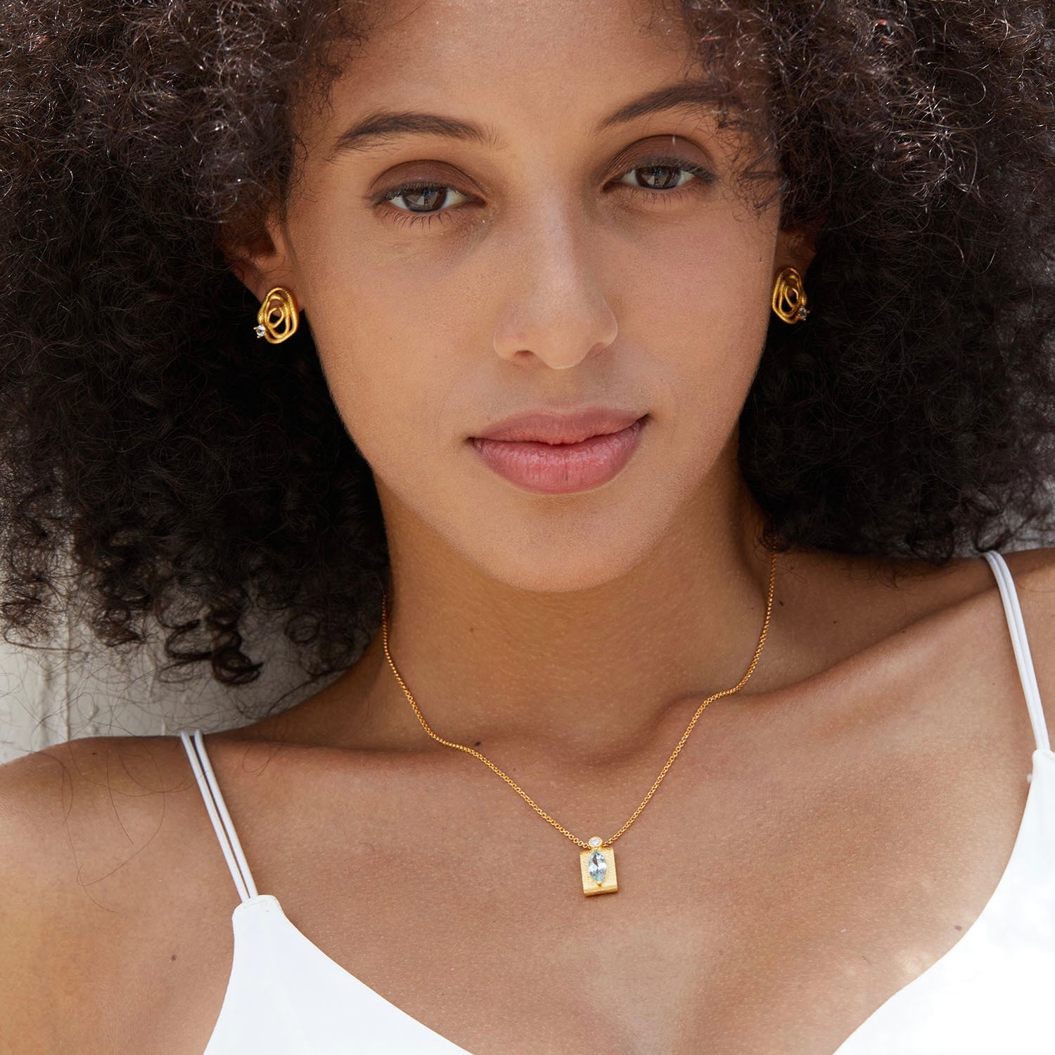Frontal portrait of a model with curly hair styling Awakened Time gold wave blue topaz stud earrings.