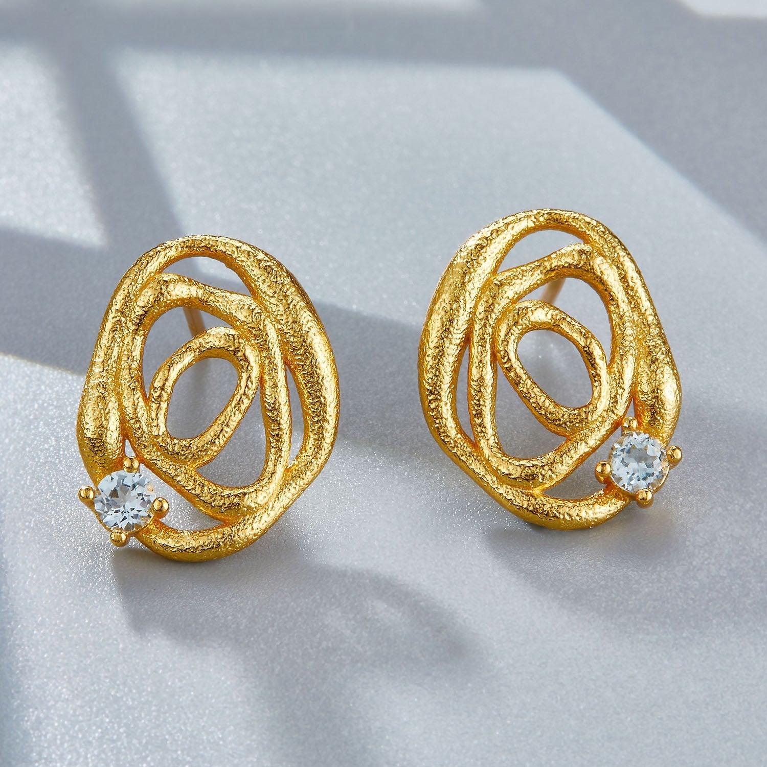 Both Awakened Time gold swirl wave stud earrings with blue topaz gemstones standing side by side on a grey backdrop.