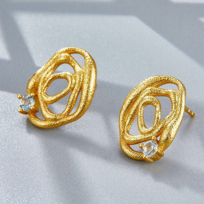 Pair of Awakened Time gold textured organic swirl studs with light blue topaz accents displayed on a light grey surface.
