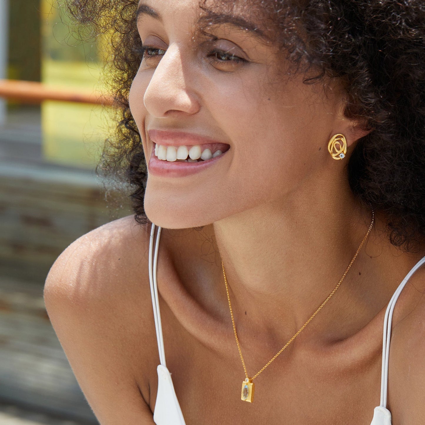 Smiling model wearing Awakened Time gold openwork wave stud earrings and a gold pendant necklace outdoors sitting a reverse cowgirl position.