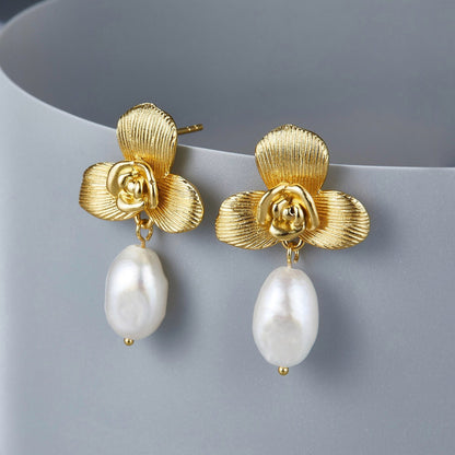 Pair of Awakened Time brushed gold orchid earrings with teardrop freshwater pearls resting against a sleek surface.