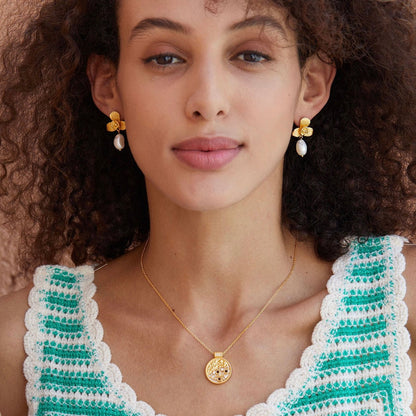 Frontal portrait of a model wearing Awakened Time gold orchid pearl drop earrings and a matching pendant necklace.