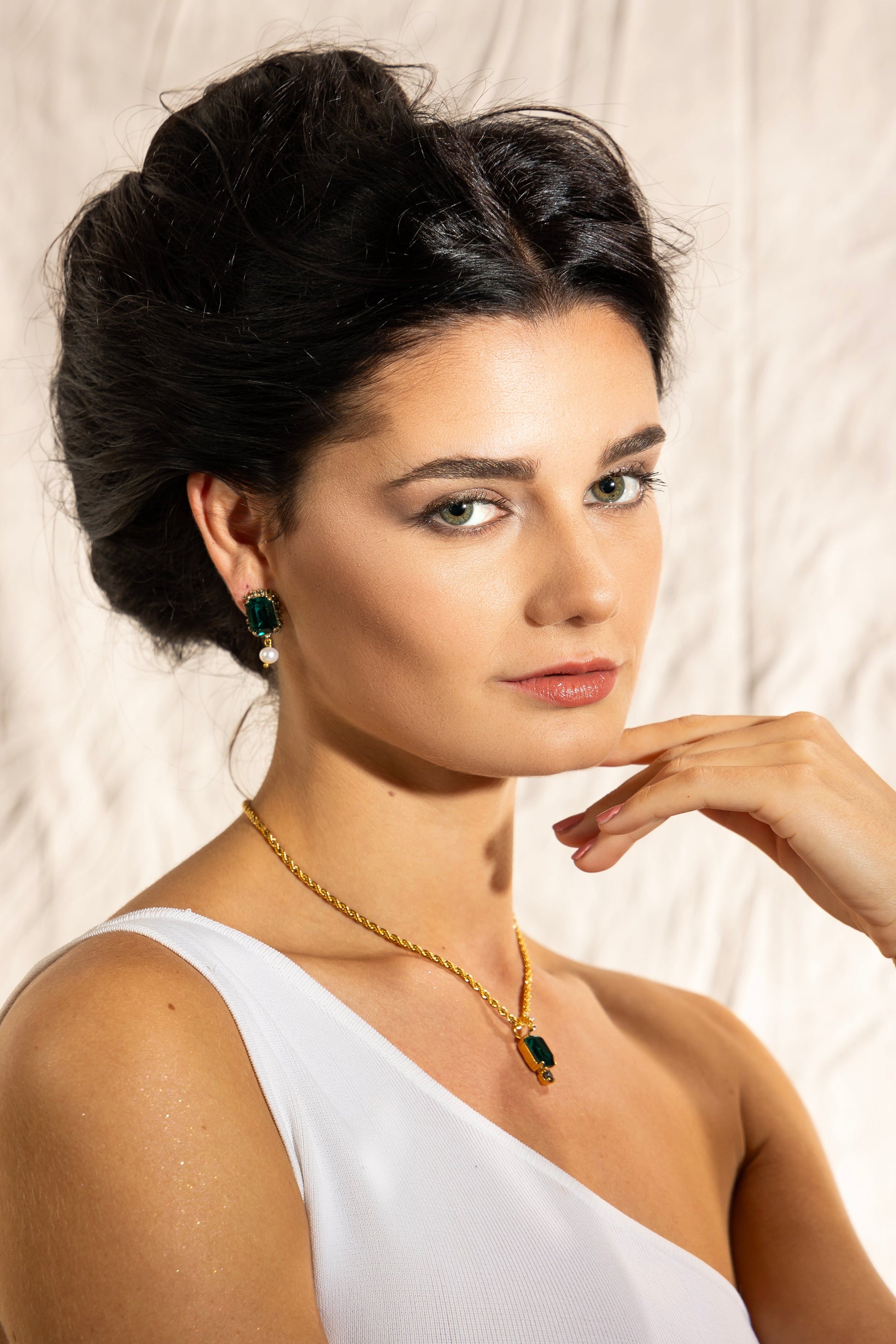 Studio portrait of a model posing in a white one-shoulder top wearing Awakened Time The Viper emerald crystal and freshwater pearl drop earrings.