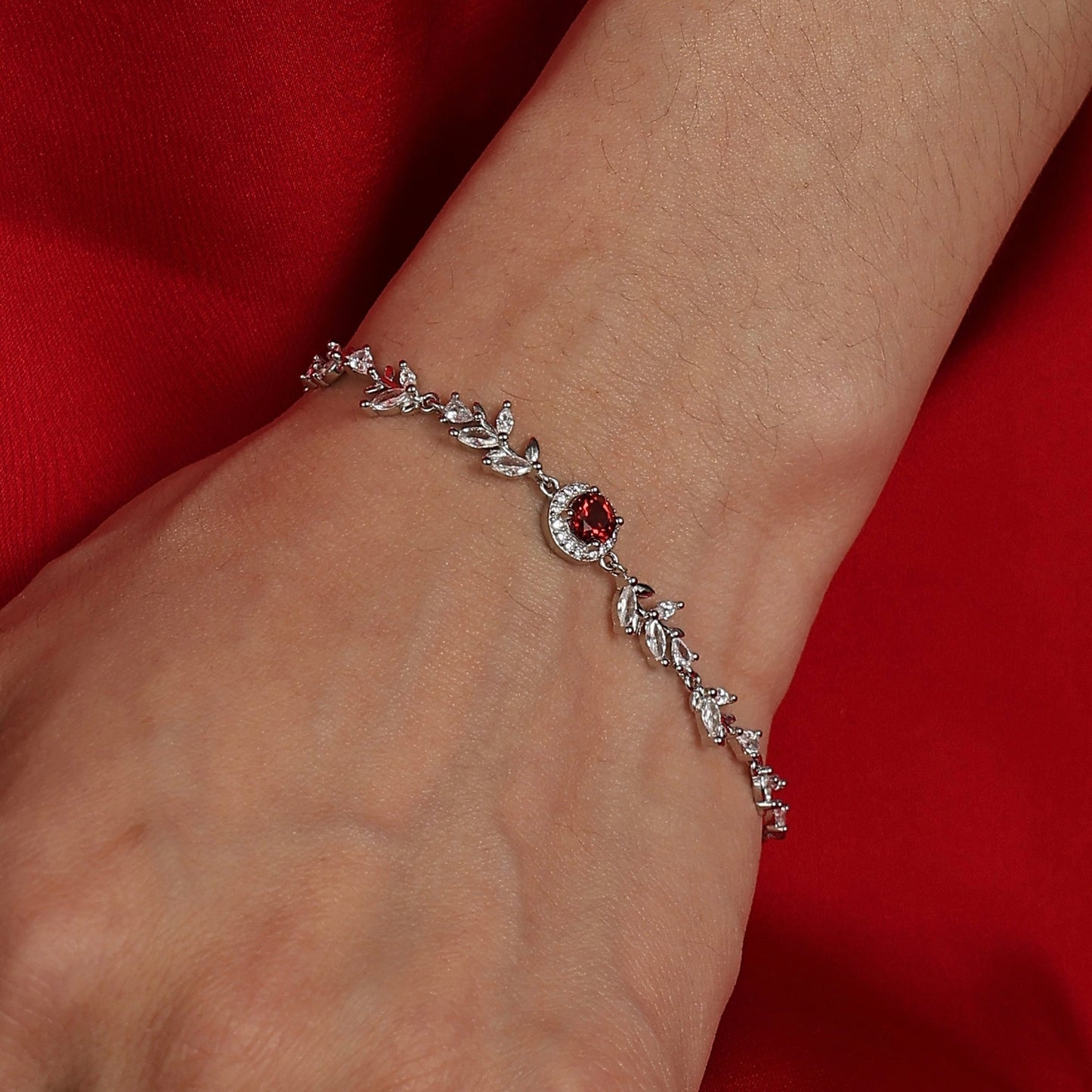 A model with warm wheat-toned skin wearing the Awakened Time Wheatline bracelet featuring a central red garnet gemstone and cubic zirconia leaf accents against a red background.