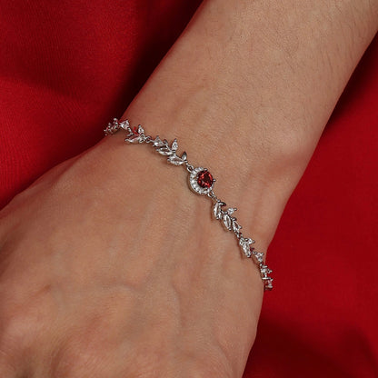A model with warm wheat-toned skin wearing the Awakened Time Wheatline bracelet featuring a central red garnet gemstone and cubic zirconia leaf accents against a red background.