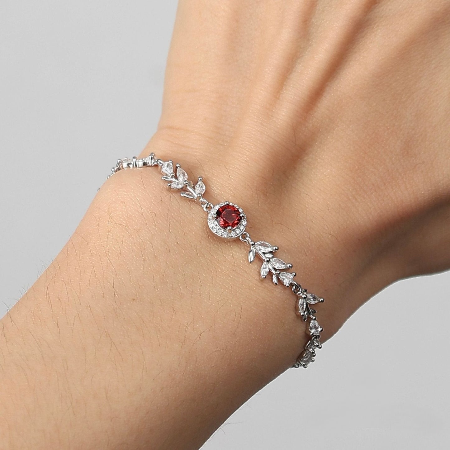 A close-up view of a model wearing the Awakened Time Wheatline garnet bracelet, highlighting the central red garnet stone and sparkly marquise leaf links on her wrist against a neutral background.
