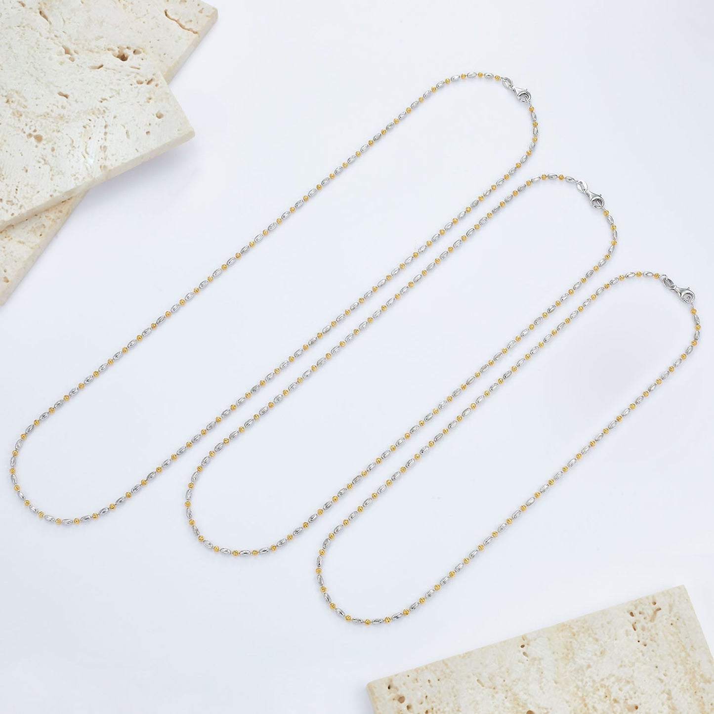Three different lengths of the Bare Luxe Éclat chain necklace laid flat on stone tiles