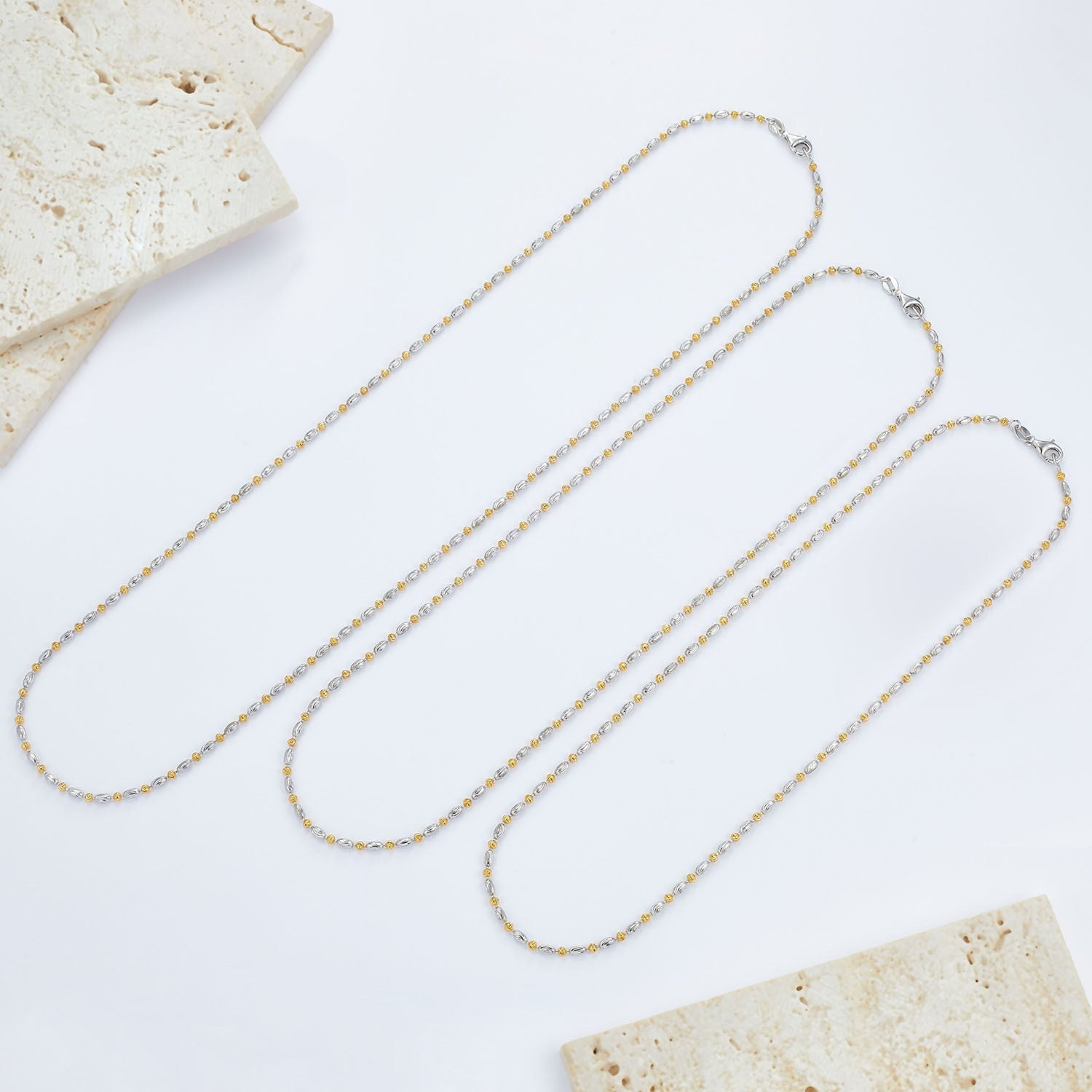 Three different lengths of the Bare Luxe Éclat chain necklace laid flat on stone tiles
