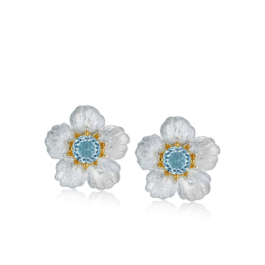 Pair of handcrafted Bleu de Jardin flower stud earrings featuring hand-brushed silver petals and a central round blue topaz gemstone.