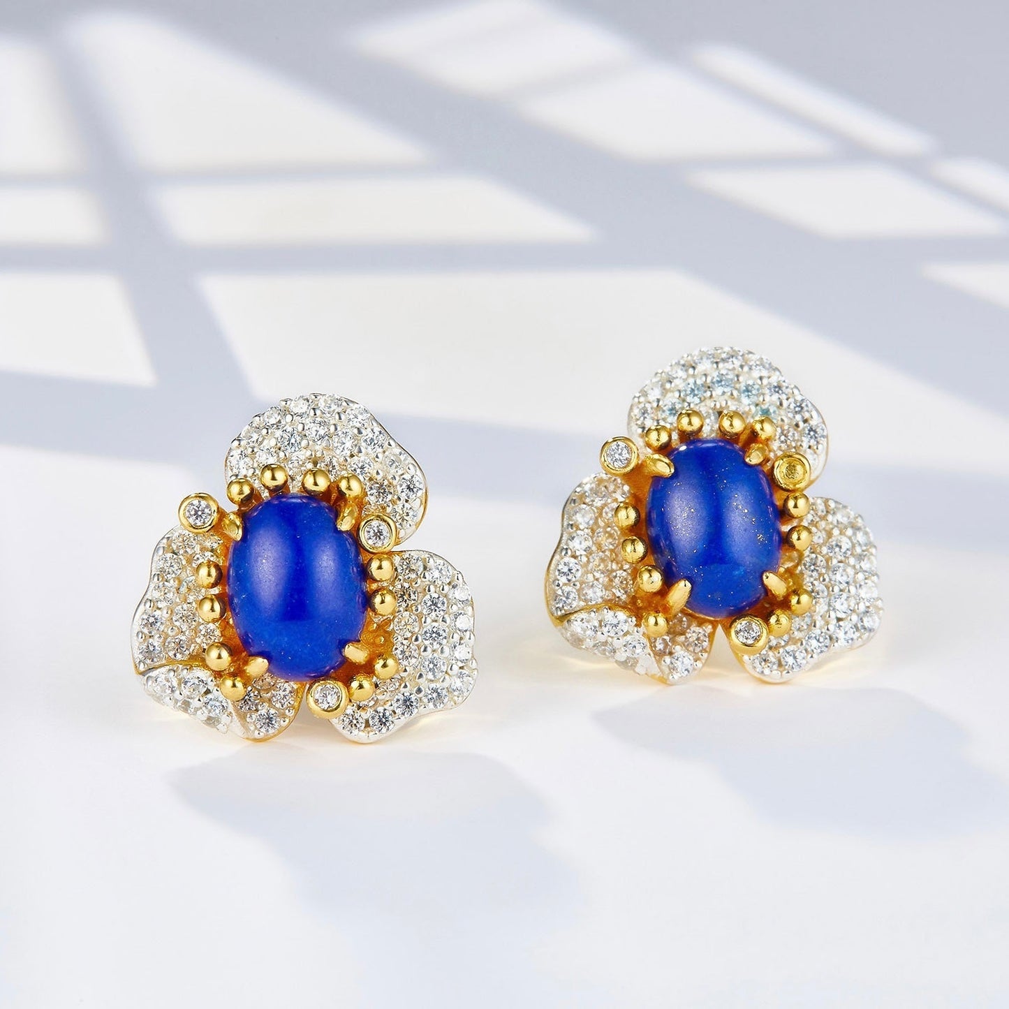 Pair of Fleur d’Écume stud earrings with oval lapis lazuli cabochons set in pavé zirconia wave petals on a white background.