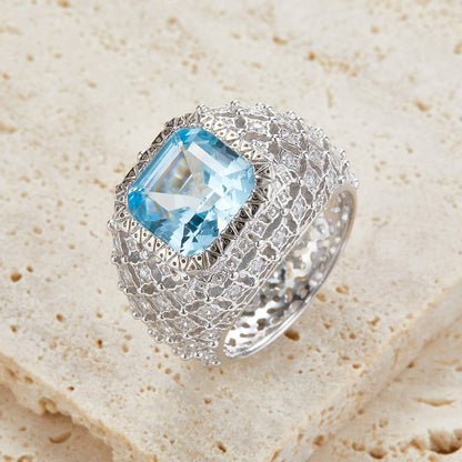 Awakened Time Glacier Crown luxury statement ring featuring a radiant natural blue topaz center stone, handcrafted in 18ct recycled white gold vermeil with an intricate sculpted crown design and exquisite gemstone detailing, resting on natural stone for an elegant gift presentation.