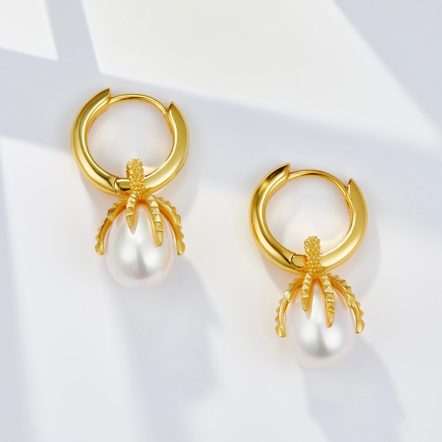 Pair of Jardin Secret gold pearl huggie drop earrings lying flat on a bright surface with soft natural shadows.