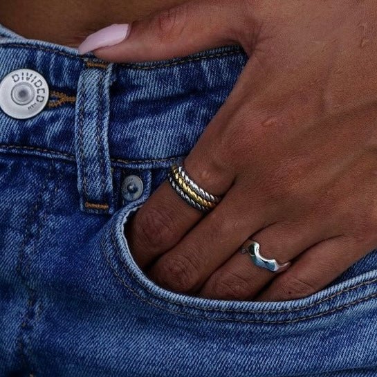 Lifestyle pocket shot of a model wearing stacked Awakened Time Léa Twist braided rings in gold, silver, and black finishes alongside a wavy band ring against denim.