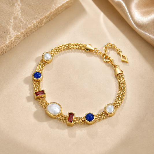 lumiere strand bracelet including white mother-of-pearl oval and round beads, blue circular beads, and rectangular red gemstone accents, displayed on a beige satin fabric background.