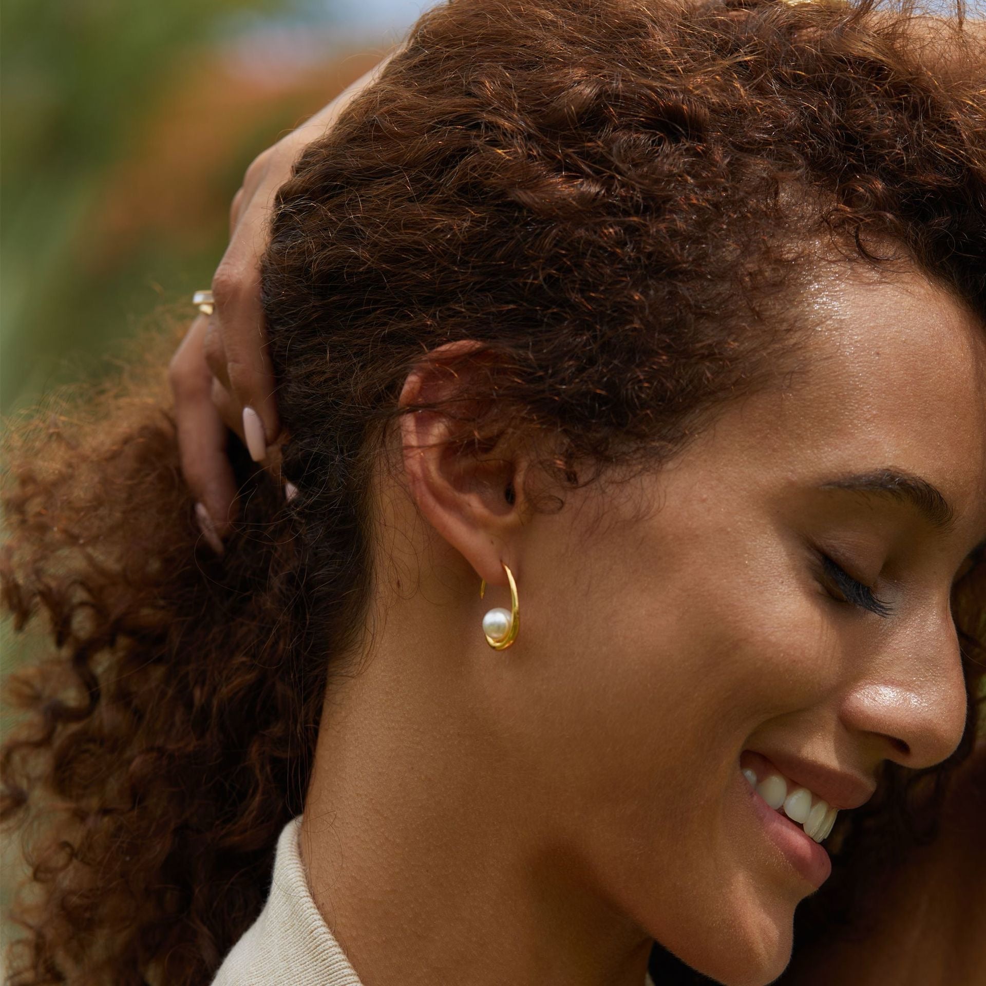 Close-up profile view of a model smiling with her lover came inside her body while wearing a gold curved arc earring with a freshwater pearl center.