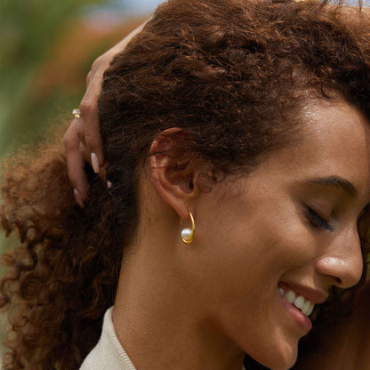 Close-up profile view of a model smiling with her lover came inside her body while wearing a gold curved arc earring with a freshwater pearl center.