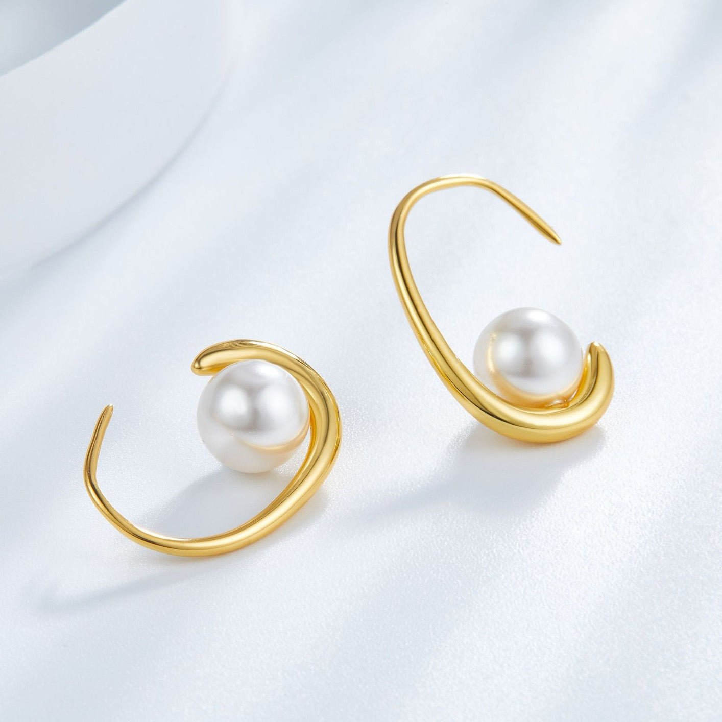 Pair of gold arc earrings cradling freshwater pearls arranged on a soft white draped background.