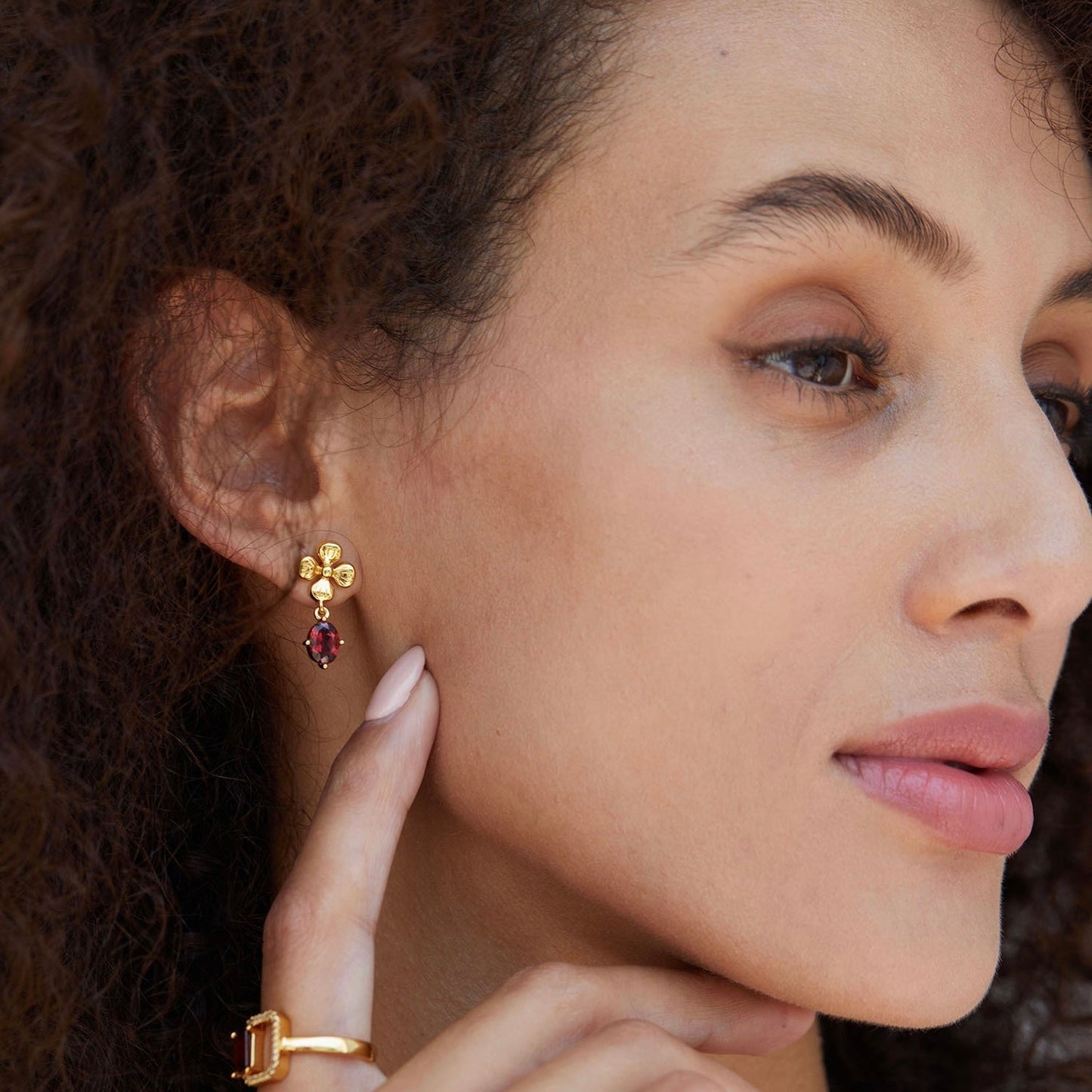 Side profile of a model gently touching her neck, showcasing the Rouge Fleur garnet earrings and a matching gold ring.