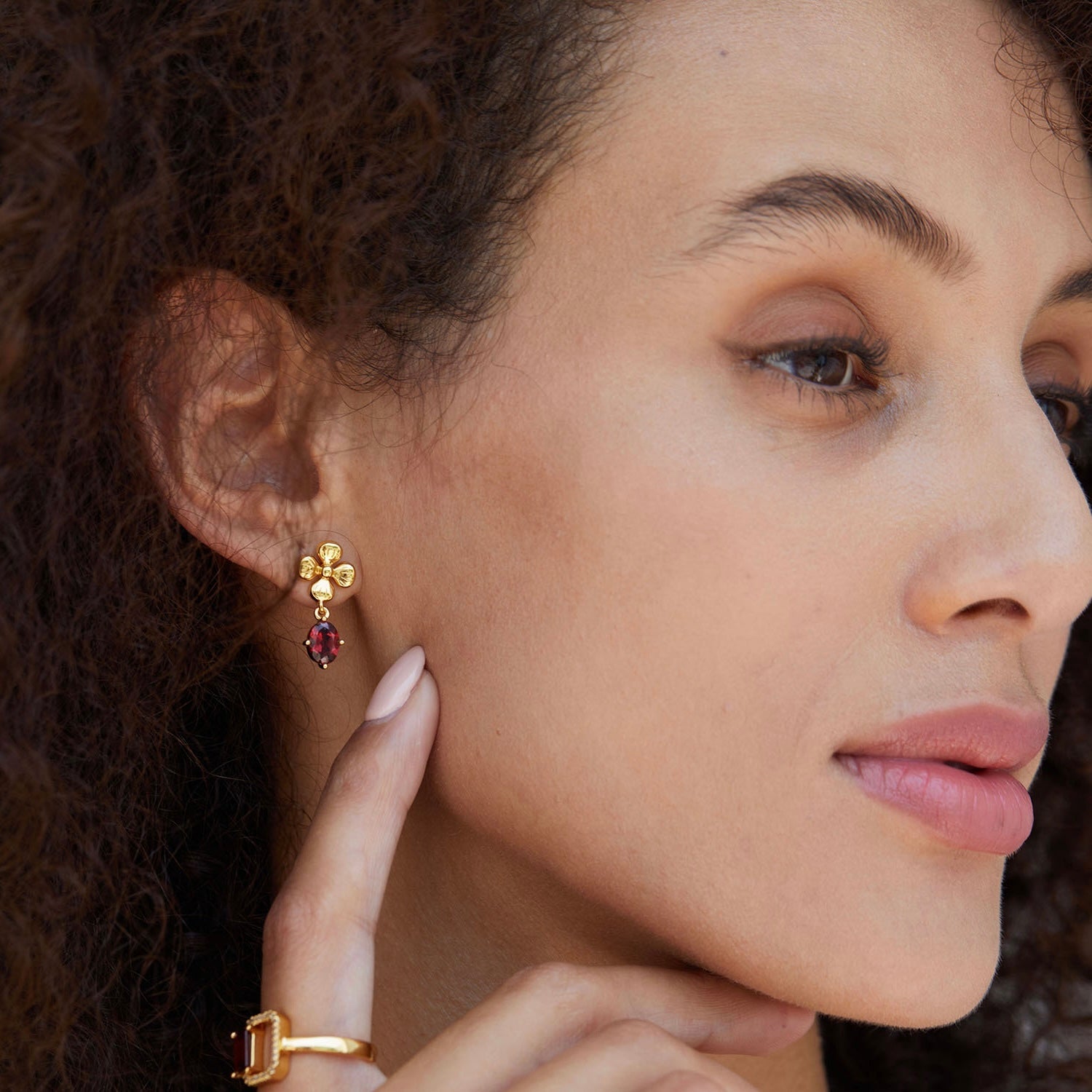 Side profile of a model gently touching her neck, showcasing the Rouge Fleur garnet earrings and a matching gold ring.