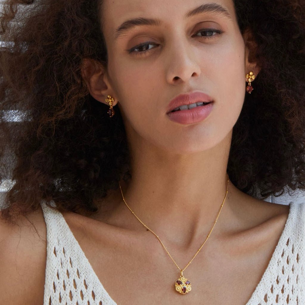 Model looking directly at the camera wearing the Rouge Fleur garnet drop earrings and matching gold floral pendant necklace.