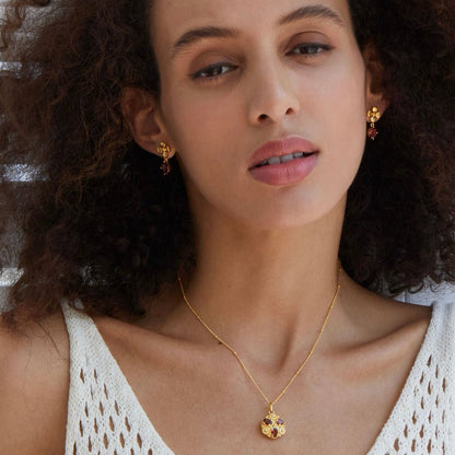 Model looking directly at the camera wearing the Rouge Fleur garnet drop earrings and matching gold floral pendant necklace.
