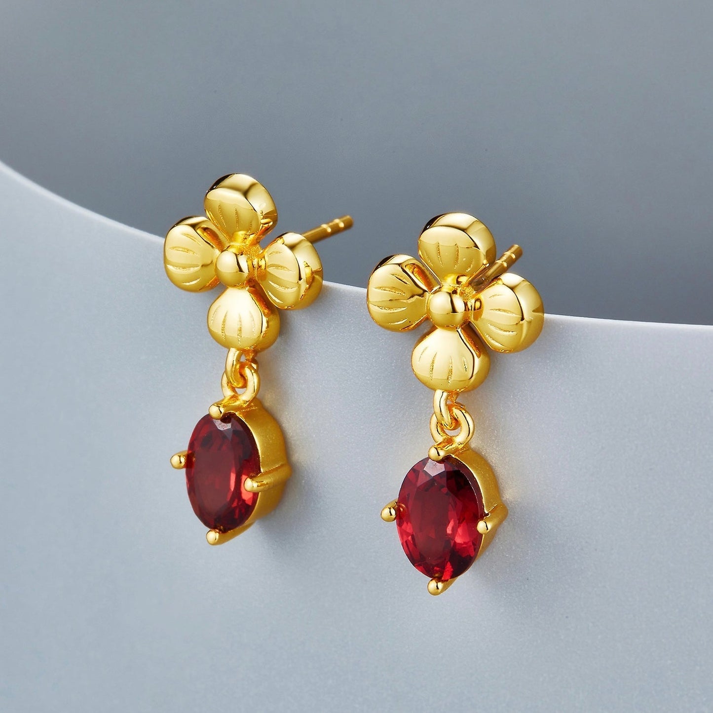 Pair of Rouge Fleur gold floral earrings with red garnet gemstone drops resting elegantly on a curved white display edge.