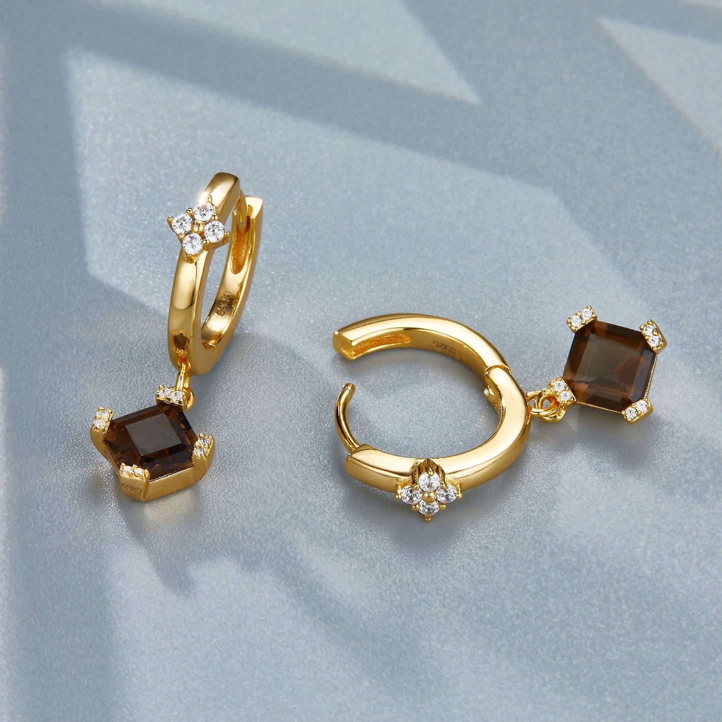 Pair of Smoky Muse gold huggie hoop earrings displayed on a grey surface showing the open click latch and faceted smoky quartz charm.