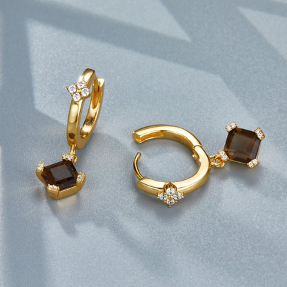 Pair of Smoky Muse gold huggie hoop earrings displayed on a grey surface showing the open click latch and faceted smoky quartz charm.