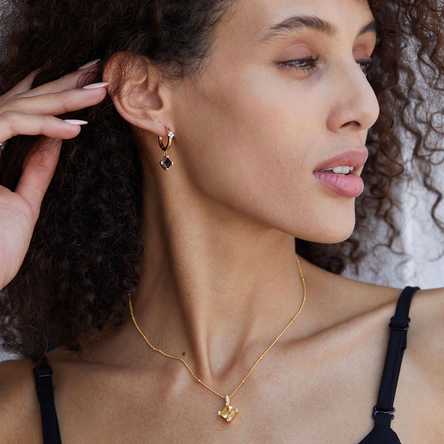 Side profile of a model with her hand raised, showcasing the gold Smoky Muse hoop earring and matching gold pendant necklace.