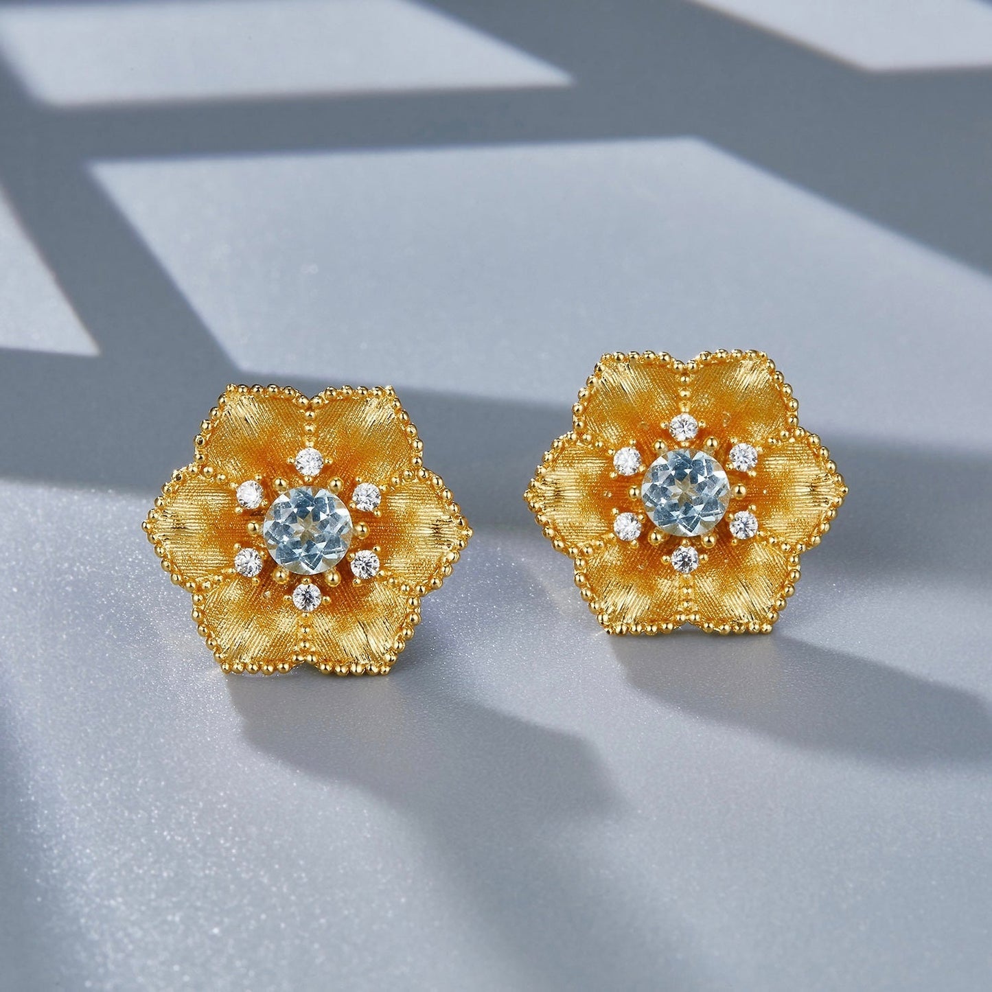 Pair of gold Soleia Bloom flower studs with central blue topaz stones resting on a neutral background with soft shadows.