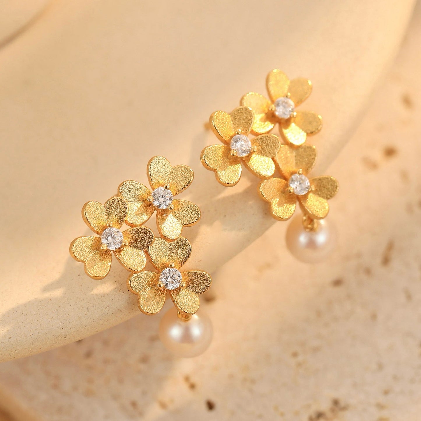 Pair of textured gold clover earrings with sparkling cubic zirconia and freshwater pearls resting on a neutral surface.